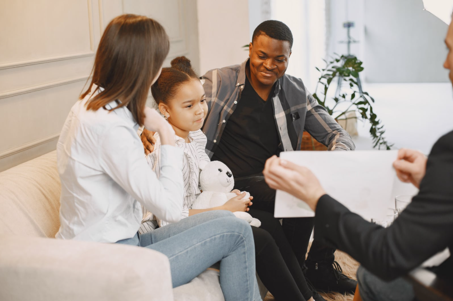 Counselor showing a paper to a family on a couch during a counseling session; a man, a woman, and a child with a stuffed bear discuss together.