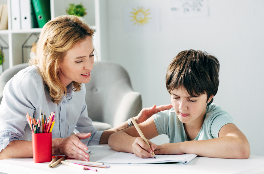 Woman helping a young boy with drawing at a desk; pencils in a red cup nearby on the table.
