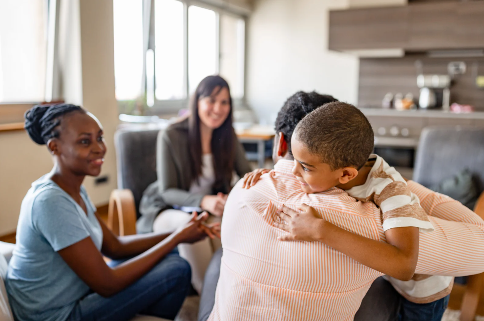 Child hugs an adult while two women chat in a bright kitchen/living area at home.
